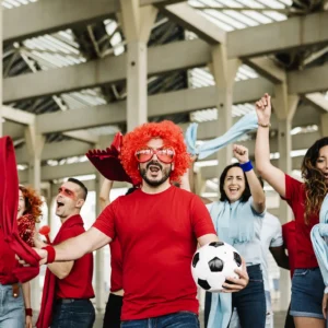 Grupo de aficionados animando con entusiasmo, vestidos con ropa temática y accesorios deportivos, representando la pasión y conexión emocional entre fans y el deporte.