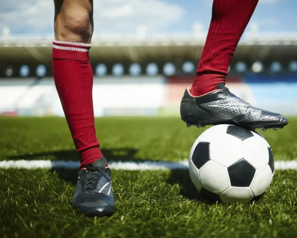 Piernas de jugadores de fútbol en un campo de césped, con un balón bajo uno de los pies, simbolizando el dinamismo y la conexión del deporte.